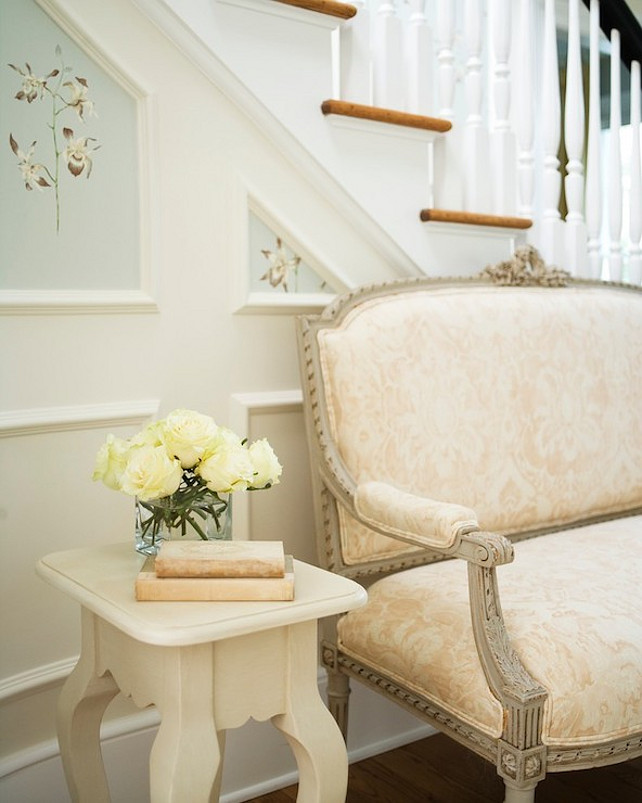 Entryway. Classic French Entryway. Elegant entryway with French gray settee upholstered in a blush pink damask fabric with an ivory cabriole leg side table to the left topped with ivory roses framed by wainscoting paneled staircase wall with handpainted floral panels.  #Entryway #FrenchInteriors