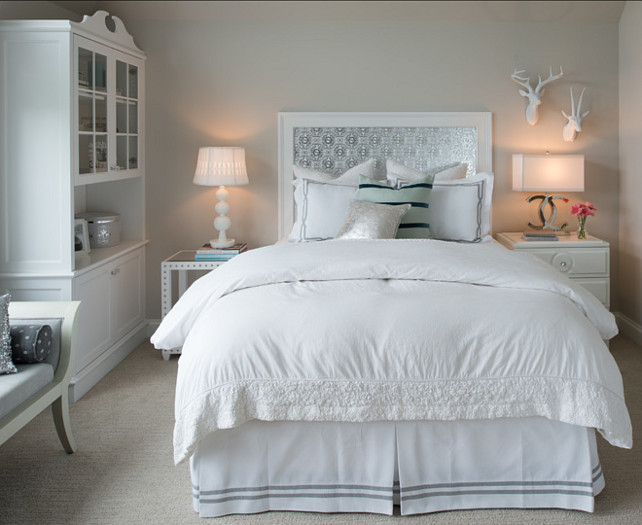 Neutral Bedroom. Beautiful neutral bedroom with greige wall paint color. Neutral Bedroom. Beautiful neutral bedroom with greige wall paint color. Designed by Studio M Interiors. #Bedroom #GrayBedroom #GreyBedroom #GreigeBedroom #Greige This gray and white bedroom features a custom silver patterned headboard with white frame. The bed is flanked by a white nailhead side table with a white stacked sphere lamp to the left and a 2-drawer nightstand with Chanel lamp on the right below a pair of faux taxidermy deer mounts. The bedroom is finished with a white glass topped hutch to the left, beside an ivory and gray bench, with figurative fashion art on the opposite wall. #Bedroom #GrayBedroom #GreyBedroom #GreigeBedroom #Greige