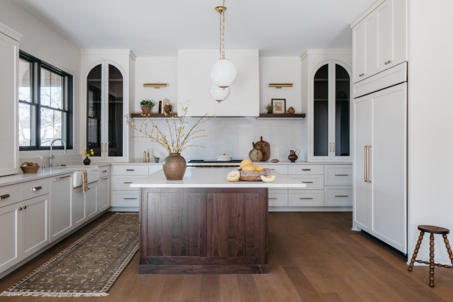 Warm White Kitchen The arched upper cabinets feature walnut interiors to match the island and shelves flaking the range hood Warm White Kitchen The arched upper cabinets feature walnut interiors to match the island and shelves flaking the range hood #WarmWhiteKitchen #WhiteKitchen #archedcabinet #walnutisland